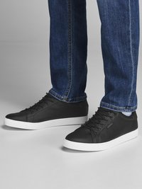 Black leather sneakers with white rubber soles, featuring round toe, lace-up front, and textured detailing on the upper. Paired with blue denim jeans.