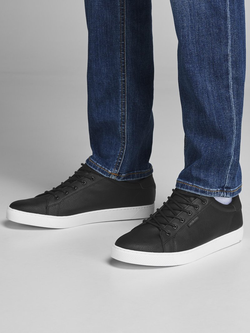 Black leather sneakers with white rubber soles, featuring round toe, lace-up front, and textured detailing on the upper. Paired with blue denim jeans.
