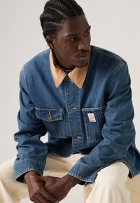 Μπλε τζιν μπουφάν με γιακά από μπεζ κορντόρι, κλείσιμο με κουμπιά και δύο τσέπες στο στήθος. Διαθέτει μια μικρή ετικέτα με το λογότυπο στην τσέπη.