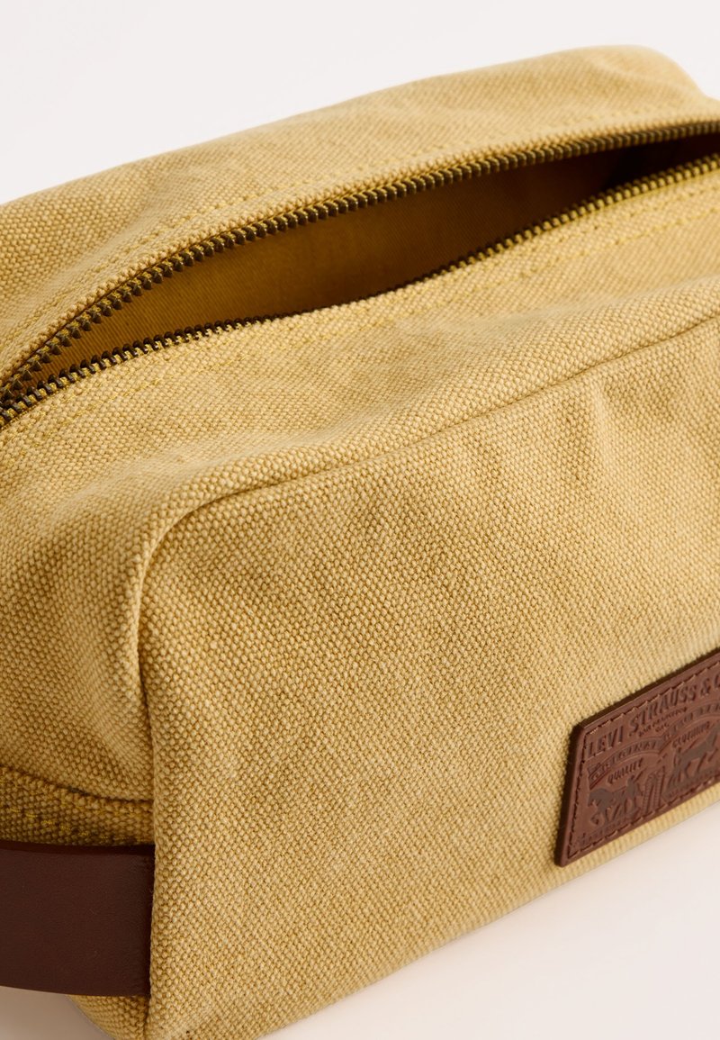 Borsa in tessuto giallo texturizzato con un accento in pelle marrone e chiusura a zip. Presenta un'etichetta con logo su un lato.