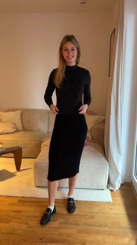 Melns garroku krekls ar baltām punktēm, kombinēts ar melnu ribotu midi svārku. Melni loaferi ar baltām zeķēm. Interjers ar dīvānu.