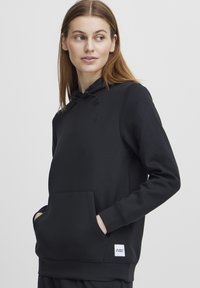Svart hettegenser med myk tekstur, frontlomme, justerbar snor i hetten, og en liten logo på brystet samt etikett på halen.