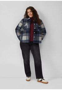 Frau mit lockigem Haar, die eine blau-grau karierte Jacke, einen burgunderfarbenen Rollkragenpullover, schwarze Jeans und weiße Turnschuhe trägt, steht vor einer schlichten Wand.