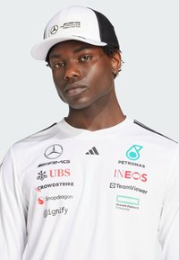 Camisa de manga larga blanca con múltiples logotipos de patrocinadores, detalles en negro, escote redondo, combinada con una gorra de malla blanca y negra que lleva un logotipo.