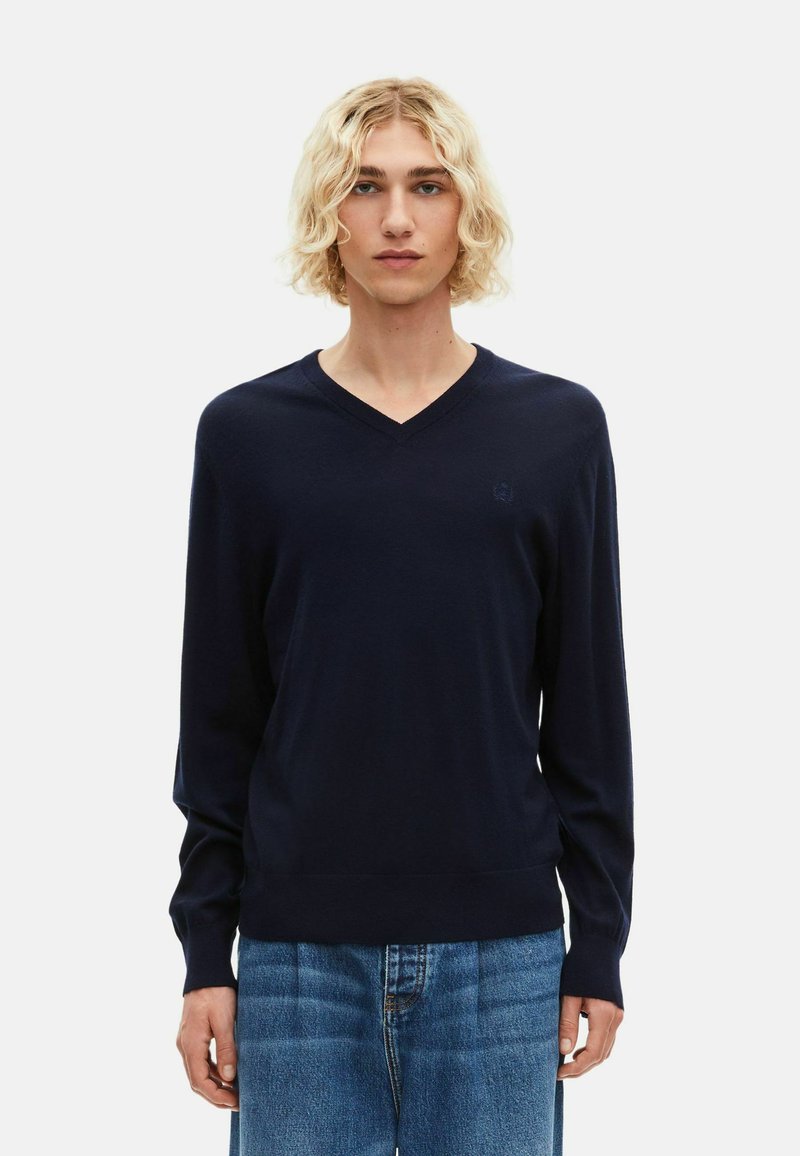 Navy-Pullover aus weichem Stoff, mit V-Ausschnitt und langen Ärmeln. Dezentes Logo-Detail auf der Brust; kombiniert mit hellblauen Jeans.