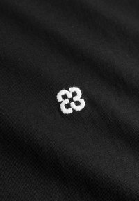 Gros plan sur un tissu noir avec un petit logo blanc brodé représentant quatre boucles entrelacées au centre.