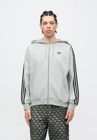 Camisola com capuz cinzenta com fecho de correr e bolso central, com mangas às riscas pretas e um pequeno logótipo preto no peito, usada com calças estampadas.