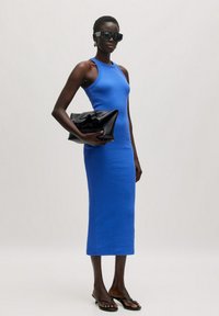 Blaues, ärmelloses Midikleid aus glattem Stoff, kombiniert mit einer schwarzen Clutch und flachen Sandalen, vor einem neutralen Hintergrund.