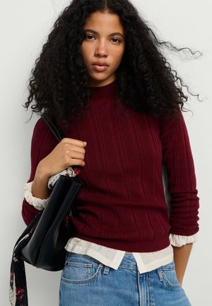 Giovane donna con capelli ricci che indossa un maglione rosso a coste sopra una camicia bianca, jeans blu e tiene una borsa nera con una sciarpa.