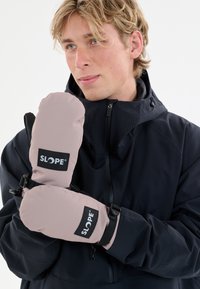Gants en tissu rose pâle avec des accents et un logo noir. Présentent des sangles ajustables et une forme arrondie. Portés par une personne en veste noire.
