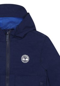 Giacca blu navy con zip frontale, cappuccio e fodera in rete. Presenta un logo bianco a forma di albero sul petto e una finitura in tessuto testurizzato.