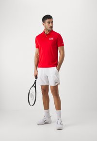 Red polo shirt with EA7 logo, white shorts with Nike tick, holding a tennis racket, and wearing white Nike shoes and socks.
