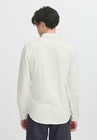 Long-sleeved shirt in off-white fabric, featuring a button-down collar, rounded hem, and textured surface detail. Back view shown.
