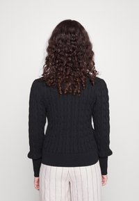 Černý svetr s kabelovým vzorem a přiléhavým střihem, s žebrovaným lemem a nabíranými dlouhými rukávy, doplněný světlými pruhovanými kalhotami.