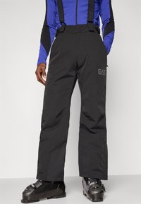 Melni slēpošanas bikses ar regulējamām piekārēm, ar šauru piegriezumu, izturīgu audumu un redzamu zīmolu uz augšstilba. Tekstūrēts apdare.