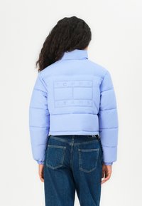 Kuttet blå puffjakke med høy krage og elastiske mansjetter, med et preget logo på ryggen, sammen med blå denimjeans.