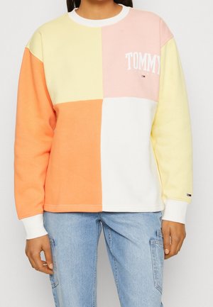Persoon draagt een colorblock-sweatshirt in pastelgeel, perzik, roze en wit met de tekst "TOMMY" en logo op de borst, gecombineerd met lichtblauwe spijkerbroek.