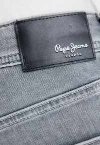 Tejido de mezclilla gris con costuras visibles, con una etiqueta de cuero negro que dice "Pepe Jeans London". Vista de cerca del área del bolsillo trasero.