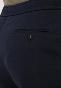 Tummansinisiä housuja, joissa on teksturoitu kangas, takatasku, koristeltu mustalla reunalla ja hopeisella napilla.