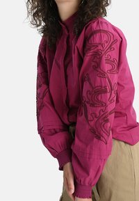 Blusa fucsia con motivo floreale ricamato, maniche a sbuffo e polsini arricciati. Presenta un colletto alla coreana e chiusura frontale con bottoni.
