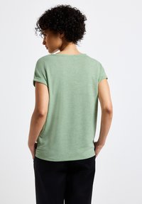 Zelené tričko s krátkým rukávem a kulatým výstřihem, vyrobené z měkké tkaniny. Má ohrnuté rukávy a volný střih, nošeno s černými kalhotami.