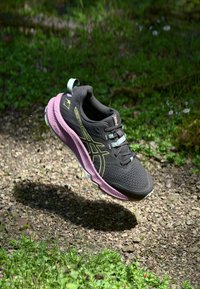 Chaussure de sport noire avec une tige texturée, une semelle intermédiaire rose et des accents verts. Comprend des lacets, un logo et une sangle de traction pour un enfilage facile.