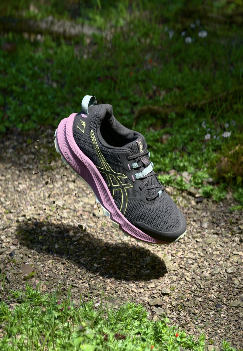 Chaussure de sport noire avec une tige texturée, une semelle intermédiaire rose et des accents verts. Comprend des lacets, un logo et une sangle de traction pour un enfilage facile.