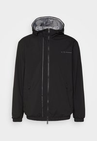 Svart jacka med huva, med ett silverfärgat foder, två blixtlåsfickor och subtil märkning i grått. Lättviktig, mjuk textur.