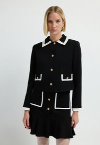 Czarna krótka kurtka i pasująca spódnica z białymi akcentami. Charakteryzuje się złotymi guzikami, kontrastowymi kieszeniami oraz rozszerzanym dołem spódnicy.