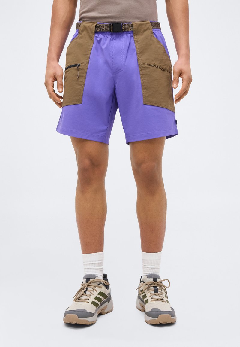 Άνδρας που φοράει μωβ σορτς με μεγάλες καφέ μπροστινές τσέπες, μπεζ και πράσινα παπούτσια πεζοπορίας και λευκές κάλτσες μέσης, στέκεται σε ουδέτερο φόντο.