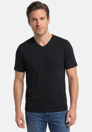 Mand stående iført en enkel sort V-hals T-shirt og blå jeans mod en ensfarvet lys baggrund, vendt fremad med et neutralt udtryk.