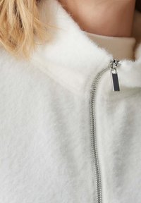 Hvid fleecejakke med høj krave, der har en sølvfarvet lynlås og glat tekstur. Nærbillede fremhæver stoffet og lynlåsdetaljen.