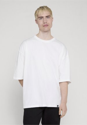 Ung person med kort blondt hår iført en stor hvid T-shirt og sorte bukser mod en ensfarvet baggrund, stående med den ene hånd bag ryggen.