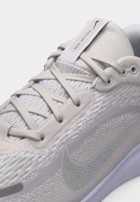 Gros plan sur une chaussure de course Nike Stellar Ride gris clair avec une tige en mesh et des lacets beige, montrant le logo Nike cousu et un col rembourré.