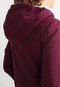 Bourgognefarvet hættejakke med tekstureret stof, synlige sømdetaljer og blødt for ved kraven for ekstra varme.