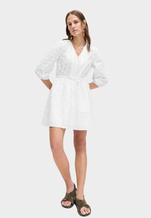 Kobieta stojąca z rękami za plecami, ubrana w białą, teksturowaną sukienkę z dekoltem w serek i rękawami 3/4 oraz oliwkowozielone sandały na otwartym palcu.