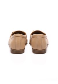 Mocasines de cuero beige con un bajo tacón de madera, que presentan una textura suave y acentos de hardware en dorado. La vista trasera muestra líneas limpias y detalles de costura.