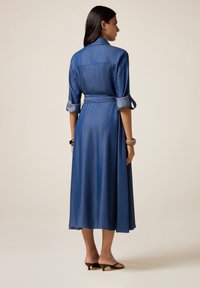 Donna con un vestito midi blu, maniche arrotolate e cintura in vita, che sta a piedi nudi su uno sfondo beige, vista da dietro.