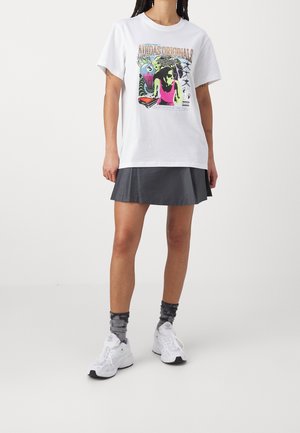 Osoba ubrana w białą koszulkę z grafiką Adidas Originals, ciemną plisowaną mini spódnicę, szare skarpetki z wzorem oraz białe trampki na jednolitym tle.