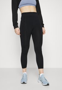 Črne hlače z visokim pasom z geometrijskimi šivi in logotipom PUMA na pasu, v paru z svetlo modrimi športnimi čevlji.