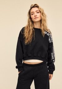 Donna con capelli biondi lunghi e mossi che indossa una felpa nera corta con testo bianco sulla spalla destra e pantaloni neri coordinati.