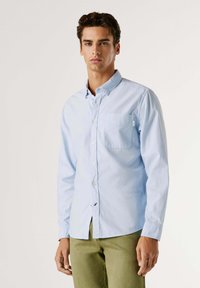 Hellblaues Langarm-Hemd mit Knopfleiste und einer Brusttasche; verfügt über einen klassischen Kragen und einen strukturierten Stoff. Kombiniert mit grünen Hosen.