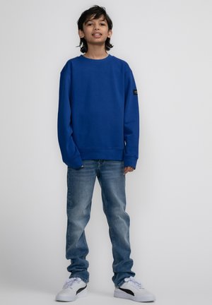Blauwe sweatshirt met lange mouwen, ronde hals en logopatch; gecombineerd met lichtblauwe jeans en witte sneakers. Tegen een effen achtergrond.