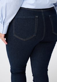 Jean bleu foncé avec une texture lisse, taille haute et deux poches arrière. Des détails de couture contrastés le long des coutures mettent en valeur le design.