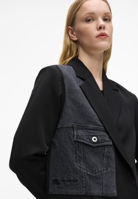 Melns žakets ar denima kreiso paneļi, kas iekļauj priekšējo kabatu, gludu tekstūru, pelēka auduma kontrastu un uzlīmētu akcentu.