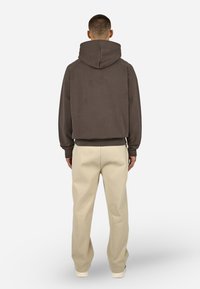 Barna kapucnis pulóver, nagyméretű hátsó kapucnival és bordázott mandzsettákkal, világos bézs, laza szabású nadrággal párosítva. Puha anyag, minimális dizájn részletekkel.