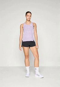 Lilla teksturert tanktop med hvite sidereflekser, parret med svarteshorts. Modellen har på seg hvite sko og sokker, og står mot en nøytral bakgrunn.