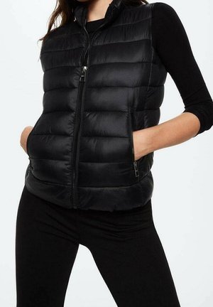 Kvinde iført en sort quiltet, ærmeløs puffer-vest over en sort langærmet top med hænderne i vestens lommer mod en ensfarvet baggrund.