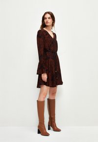 Rottöne Kleid mit schwarzem Blättermuster, langen Ärmeln, ausladenden Manschetten und einem schwarzen Gürtel. Kombiniert mit kniehohen braunen Stiefeln mit Absatz.