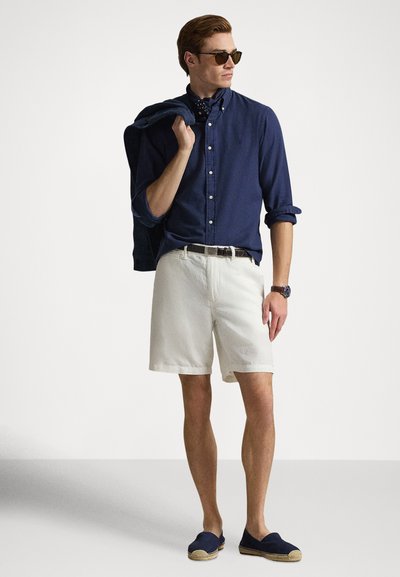 Πουκάμισο κουμπωτό ναυτικού μπλε, κρεμ σορτς, ναυτικά εσπαντρίγιες, καφέ ζώνη, γυαλιά ηλίου. Το μοντέλο κρατά ένα σακάκι, μπλε μαντήλι ναυτικού γύρω από τον λαιμό.